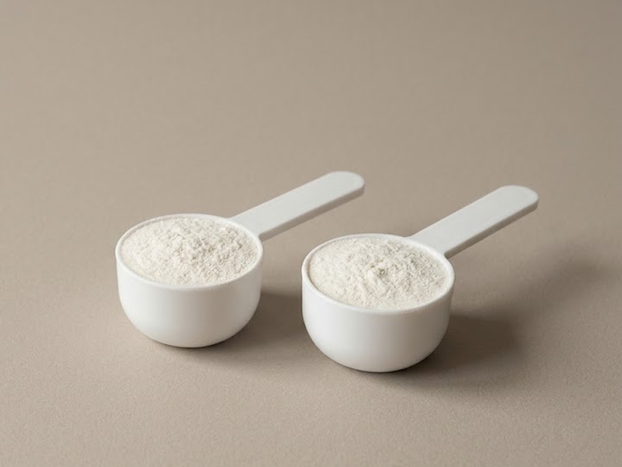 Illustration: two identical white measuring scoops, each filled level with the same fine white powder, side by side on plain taupe paper